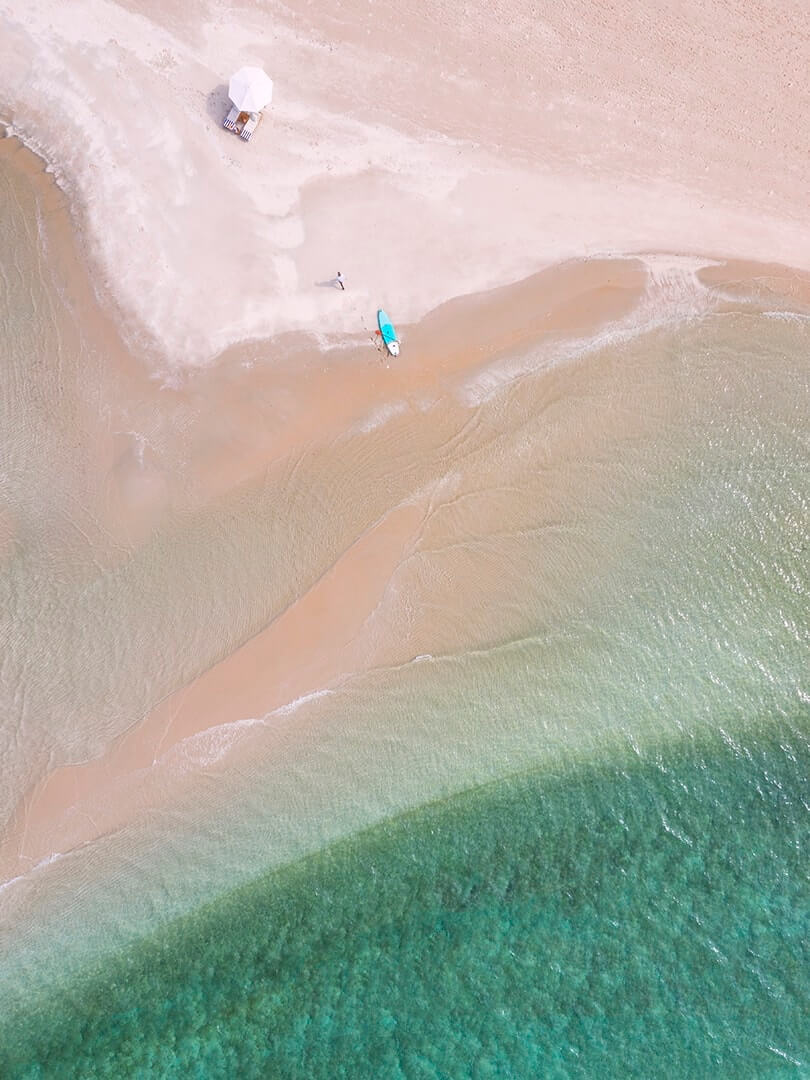 Drone shot from directly above of a paddleboard and sun loungers on a beach at Kisawa Sanctuary, Benguerra Island, photographed by Elsa Young. Drone shot from directly above of a paddleboard and sun loungers on a beach at Kisawa Sanctuary, Benguerra Island, photographed by Elsa Young.