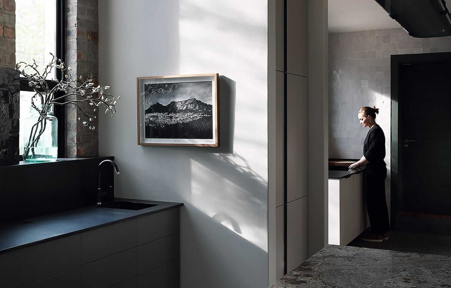 Woman in the kitchen washing up, with art of the walls and flowers on the windowsill. Photo by Elsa Young.